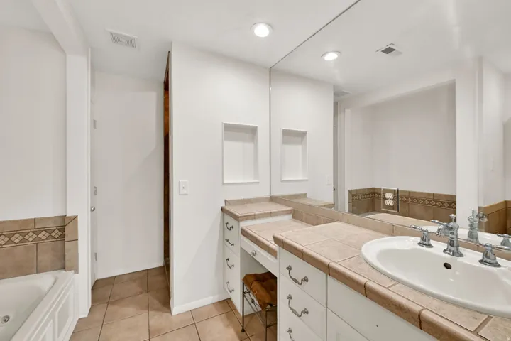 White vanity with tile countertop, integrated sink, large mirror, recessed wall shelving, and tile flooring