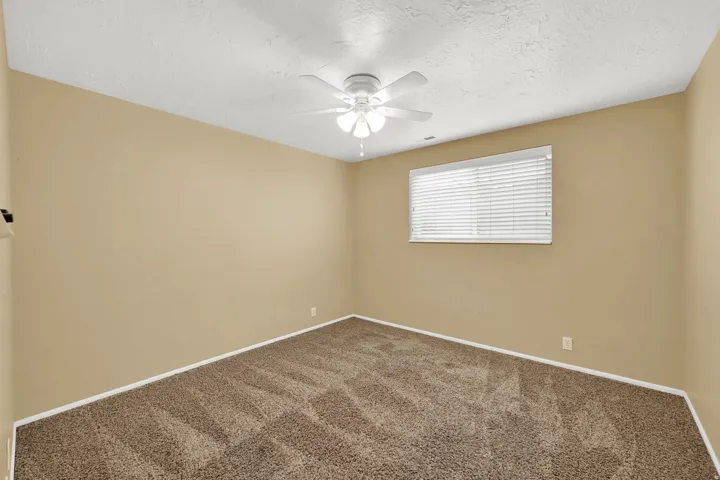 Neutral wall paint, textured ceiling, window with horizontal blinds, ceiling fan with integrated lighting, and patterned carpeting