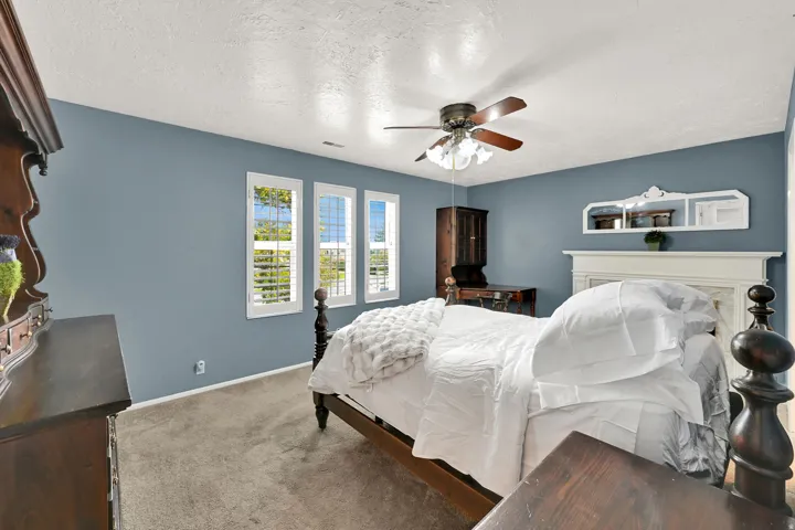 Three windows with white plantation shutters, ceiling fan with light fixture, light blue wall paint, carpet flooring, and a decorative white fireplace mantel