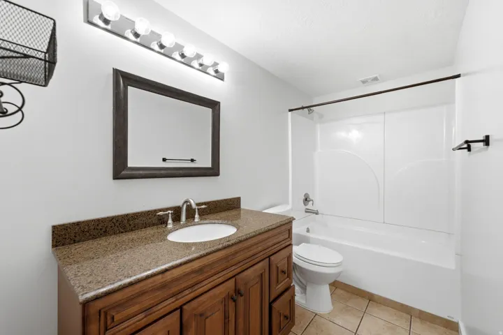 White bathtub with integrated shower, vanity with an undermount sink and a granite-finish countertop, wood-finish cabinetry, and tiled flooring