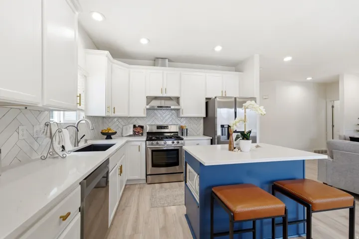 White cabinetry with gold-tone hardware, stainless steel appliances, herringbone tile backsplash, light wood-finish flooring, and a blue island with a white countertop
