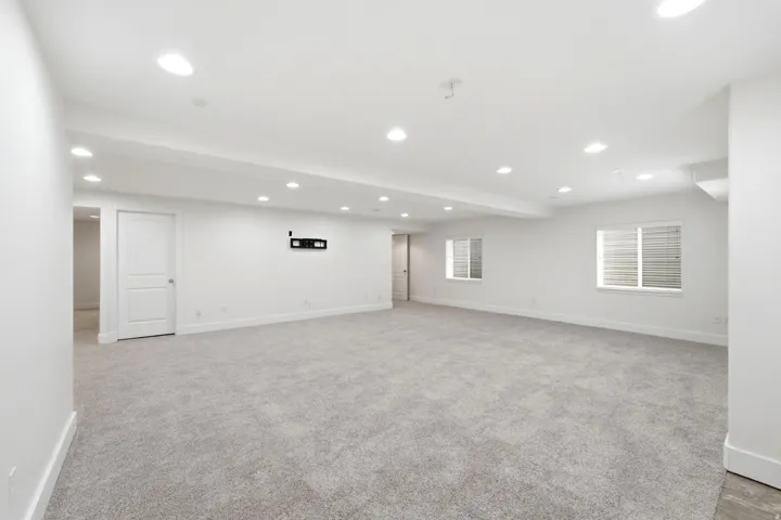 Expansive carpeted room with recessed lighting, multiple windows with horizontal blinds, and a white paneled door. Could be a game room, theatre room, oran  incredible rec room