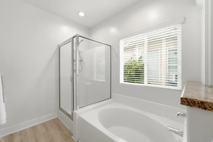 Soaking tub with chrome fixtures, glass-enclosed shower, wood-finish flooring, and a window with blinds