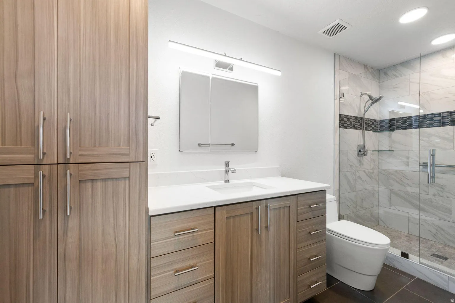 Enclosed shower with glass paneling and decorative tile band, single vanity with integrated sink and white countertop, wood-finish cabinetry with brushed nickel hardware, and built-in storage cabinets