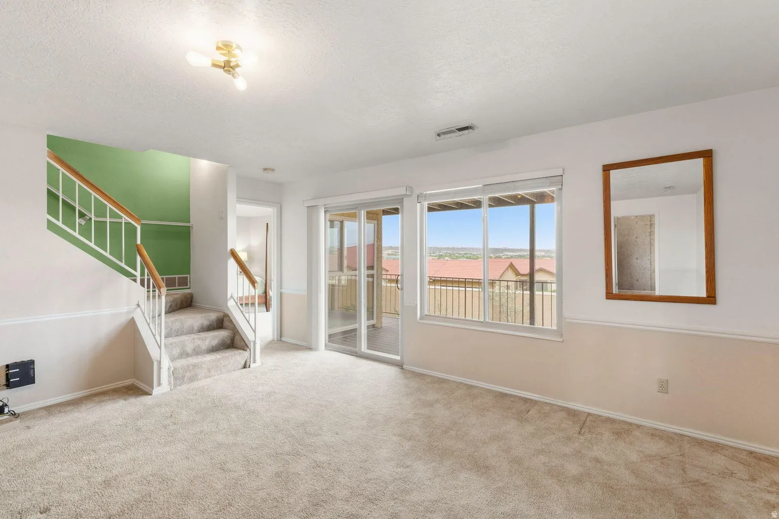 Carpeted flooring throughout, white wall paneling, staircase with white railings and wood-finish handrails, and glass sliding doors opening to an exterior balcony
