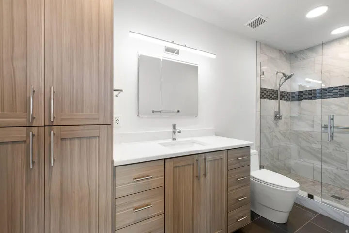 Enclosed shower with glass paneling and decorative tile band, single vanity with integrated sink and white countertop, wood-finish cabinetry with brushed nickel hardware, and built-in storage cabinets