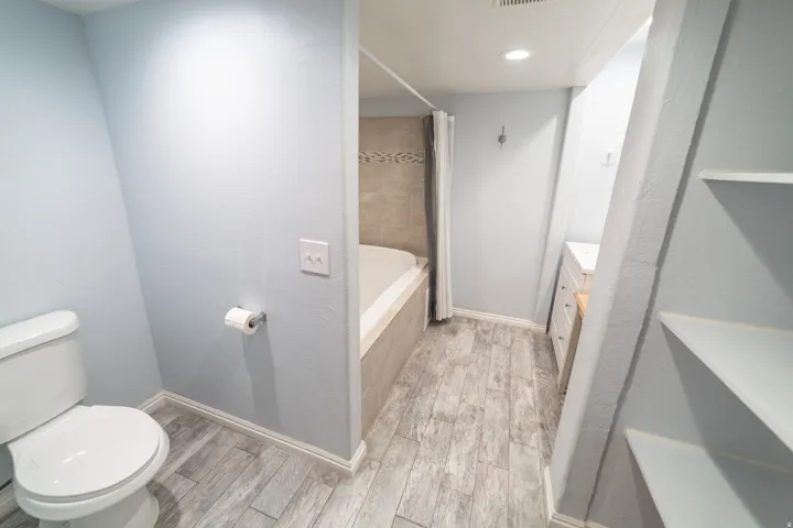 Recessed lighting illuminates wood-finish flooring, a built-in tub with tile surround, and a vanity with an undermount sink
