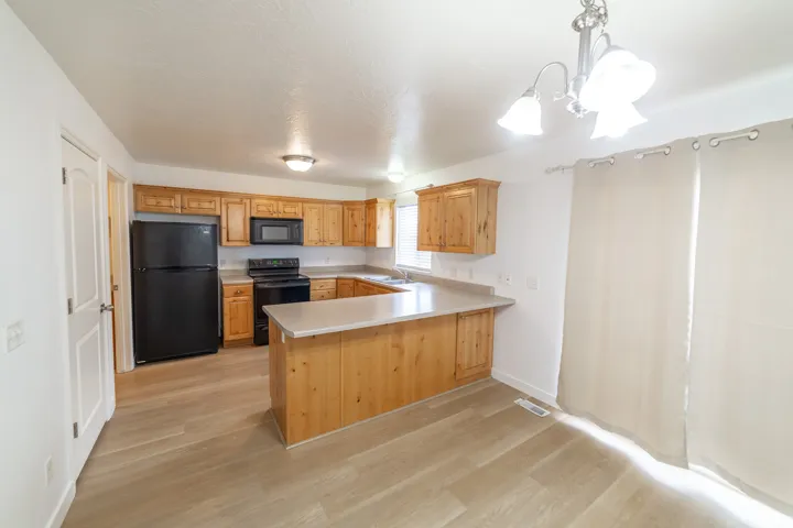 Kitchen with light wood cabinetry, L-shaped countertop, black appliances, and wood-finish flooring