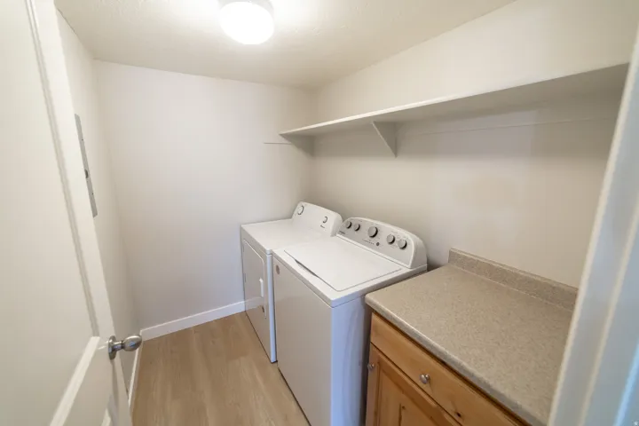 Laundry area featuring wood-finish flooring, a built-in shelf, a light-colored countertop, and light wood cabinetry with silver hardware