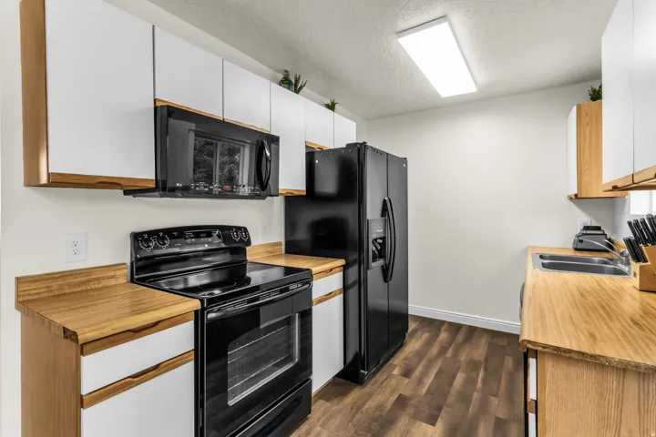 Kitchen with black appliances, white upper cabinetry, light wood-finish countertops, and wood-finish flooring