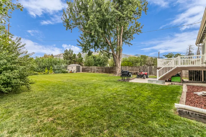 Expansive grassy lawn with mature trees, elevated wood deck with white railings, concrete patio, and a storage shed