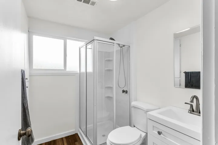 Walk-in shower with glass enclosure, white vanity with integrated sink, wood-finish flooring, and frosted window