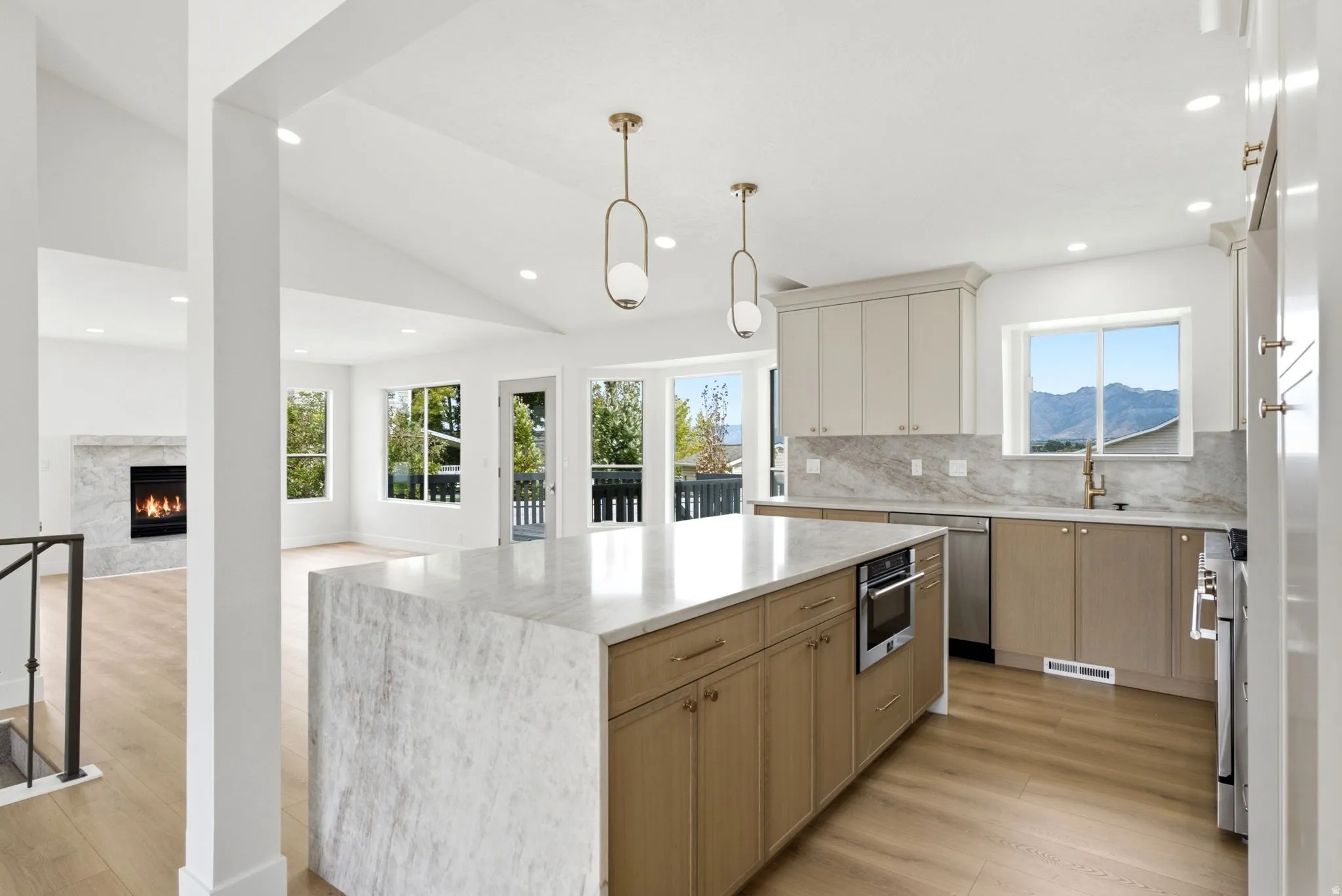 Open-concept living area featuring vaulted ceilings, wood-finish flooring, a kitchen island with built-in appliances, a stone-surround fireplace, and numerous windows