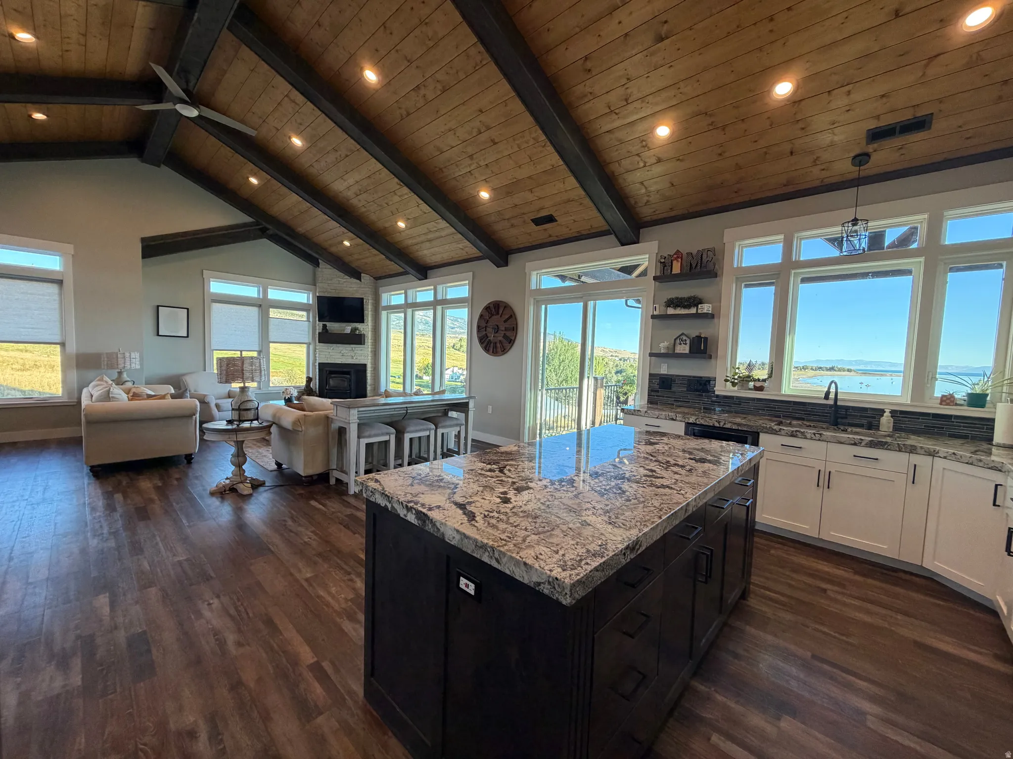 Vaulted wood-finish ceiling with exposed beams, wood-finish flooring, central kitchen island with stone countertop, white kitchen cabinetry with stone backsplash, and a fireplace with stone surround