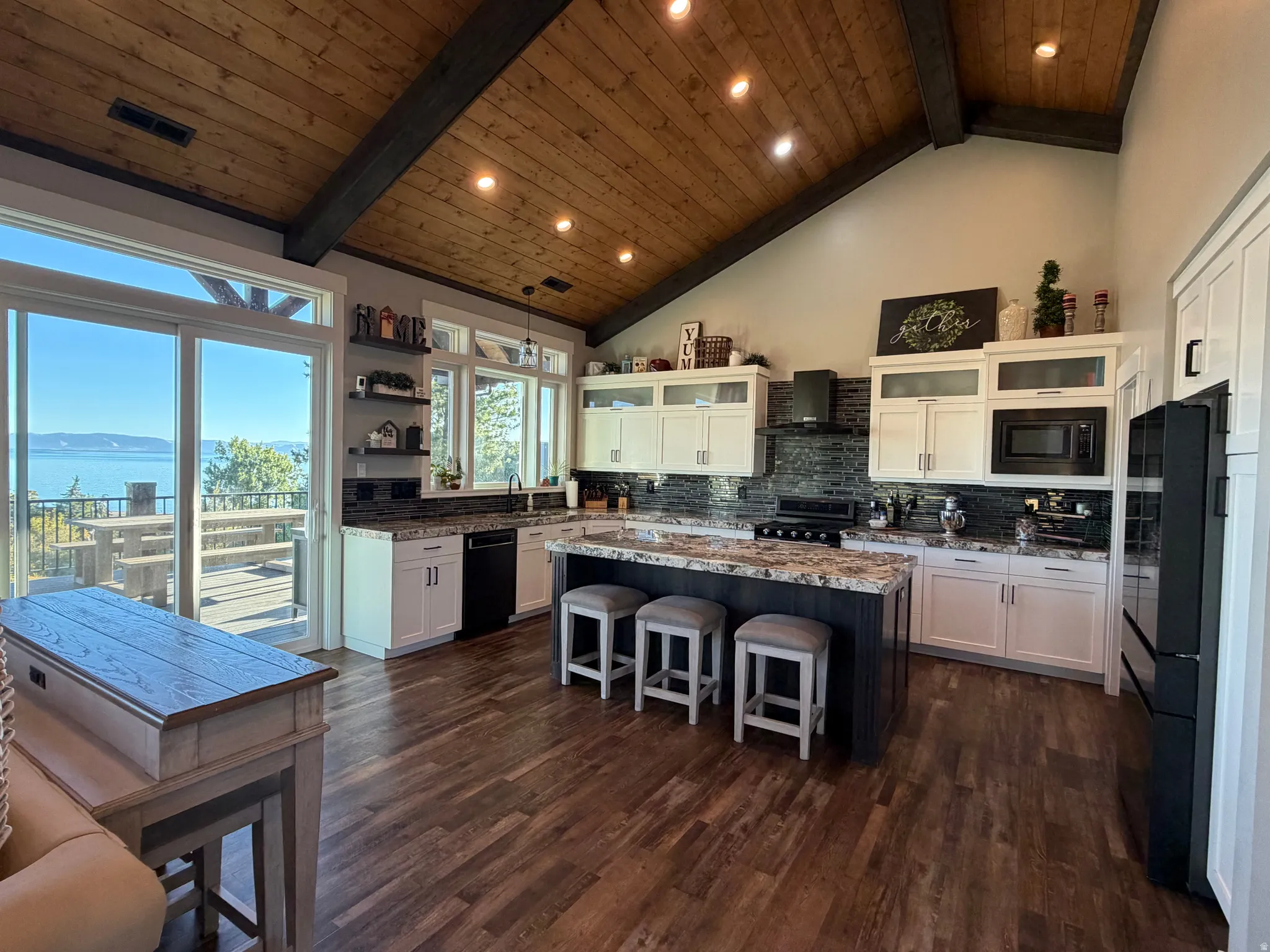 Vaulted wood-plank ceiling with exposed beams, wood-finish flooring, kitchen island with seating, white cabinetry with dark hardware, and a sliding glass door opening to an outdoor deck