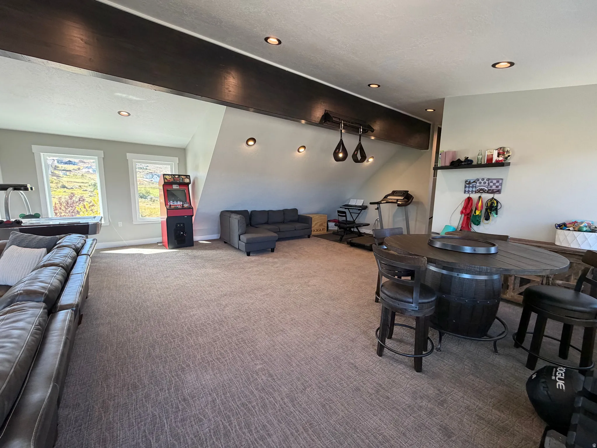 Recessed ceiling lighting, exposed wood-finish beam, two windows with white trim, wall-mounted light fixtures, and textured carpet flooring