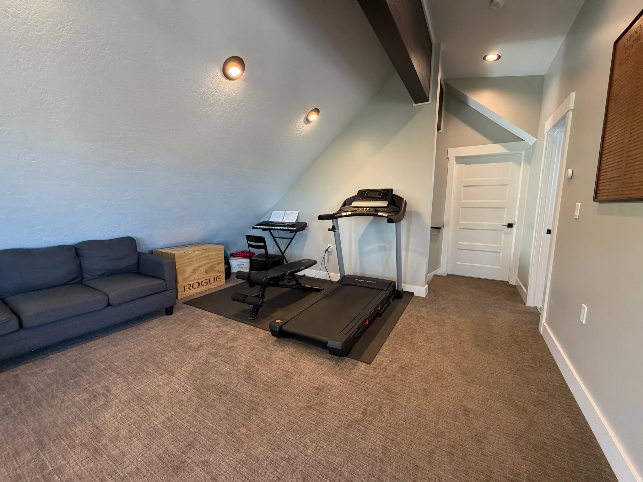 Angled ceiling with exposed wood beams, recessed wall sconces, neutral carpeting, and a white panel door