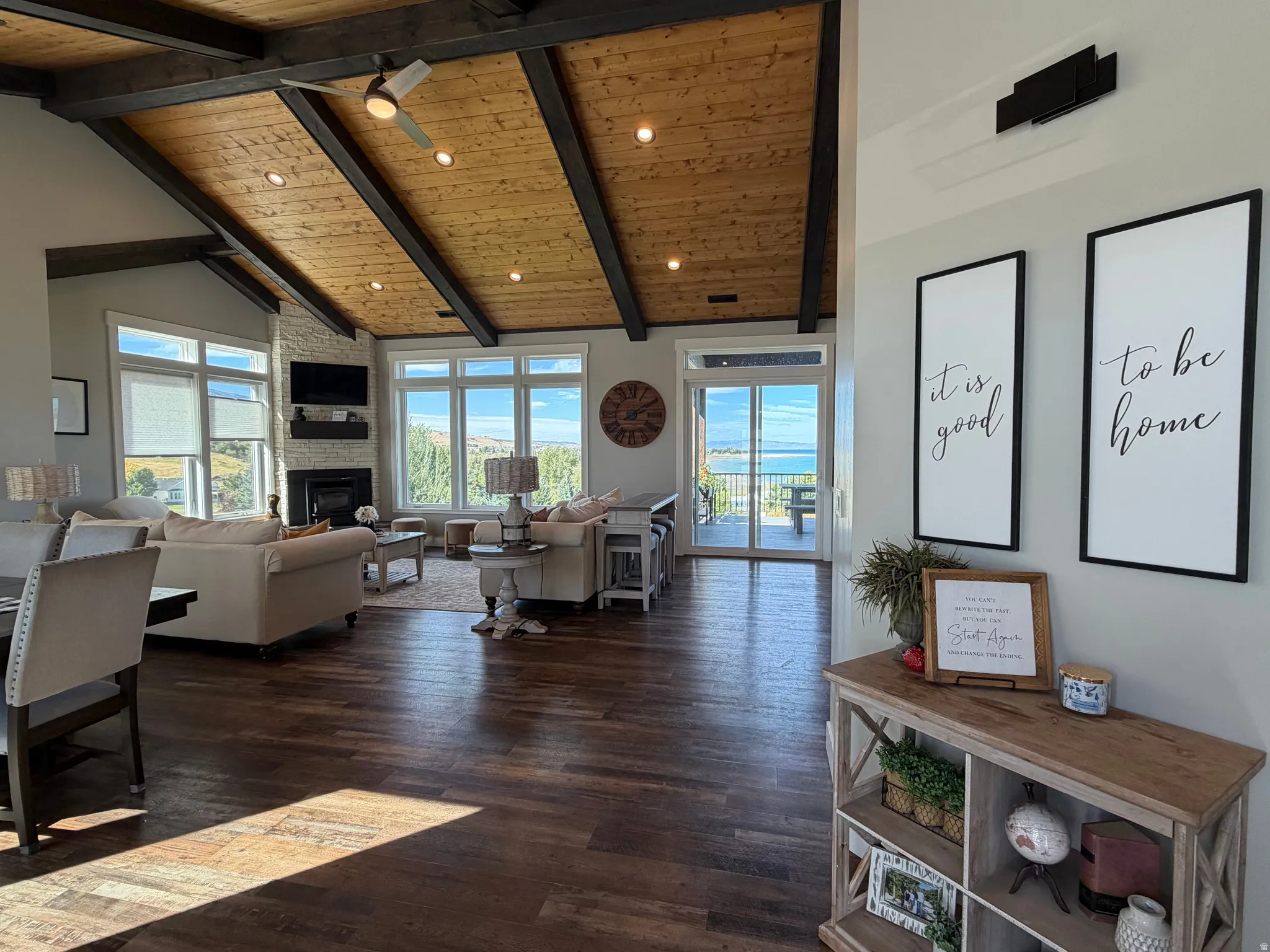 Vaulted wood-plank ceiling with exposed beams, wood-finish flooring, white brick-clad fireplace, large windows, and sliding glass doors to outdoor space