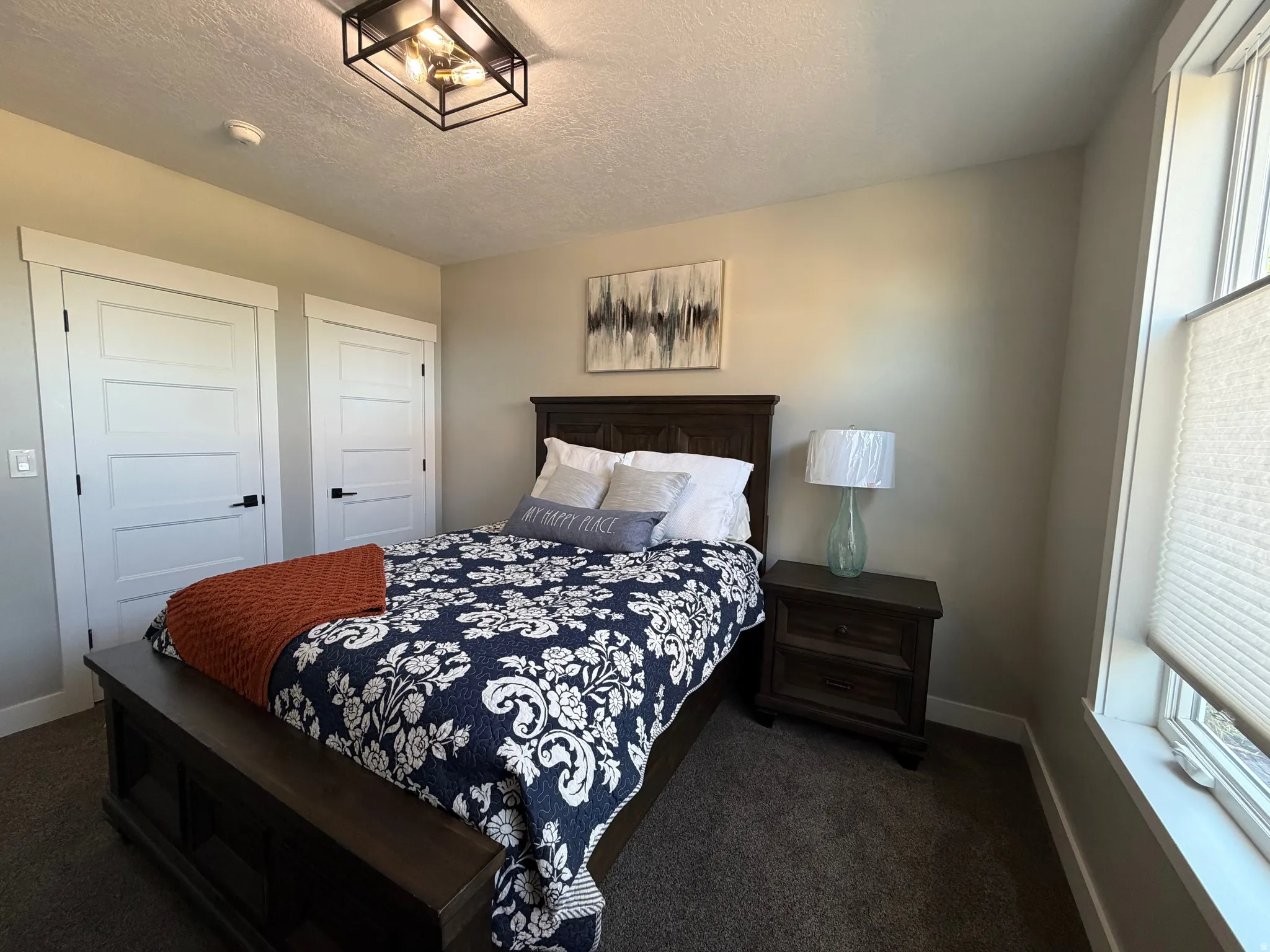 Recessed window with privacy blinds, dark wood-finish nightstand, textured carpet flooring, contemporary ceiling fixture, and modern four-panel doors