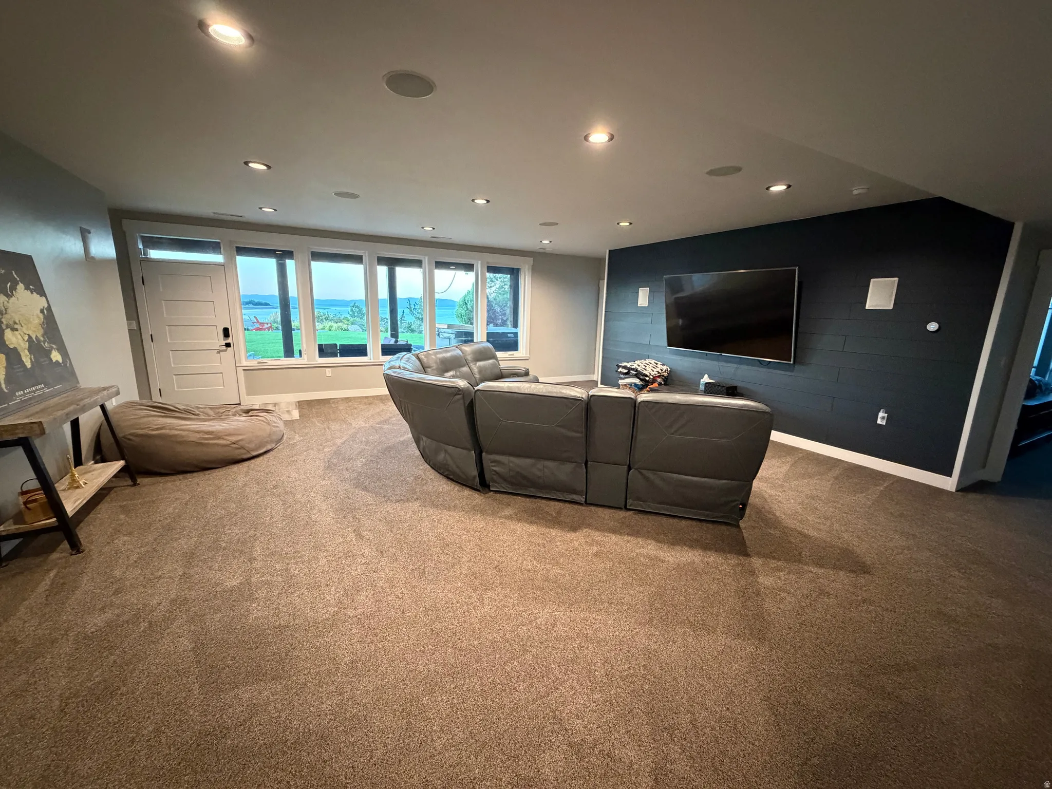 Recessed lighting, ceiling-mounted speakers, dark accent wall, carpeted flooring, and a wall of windows