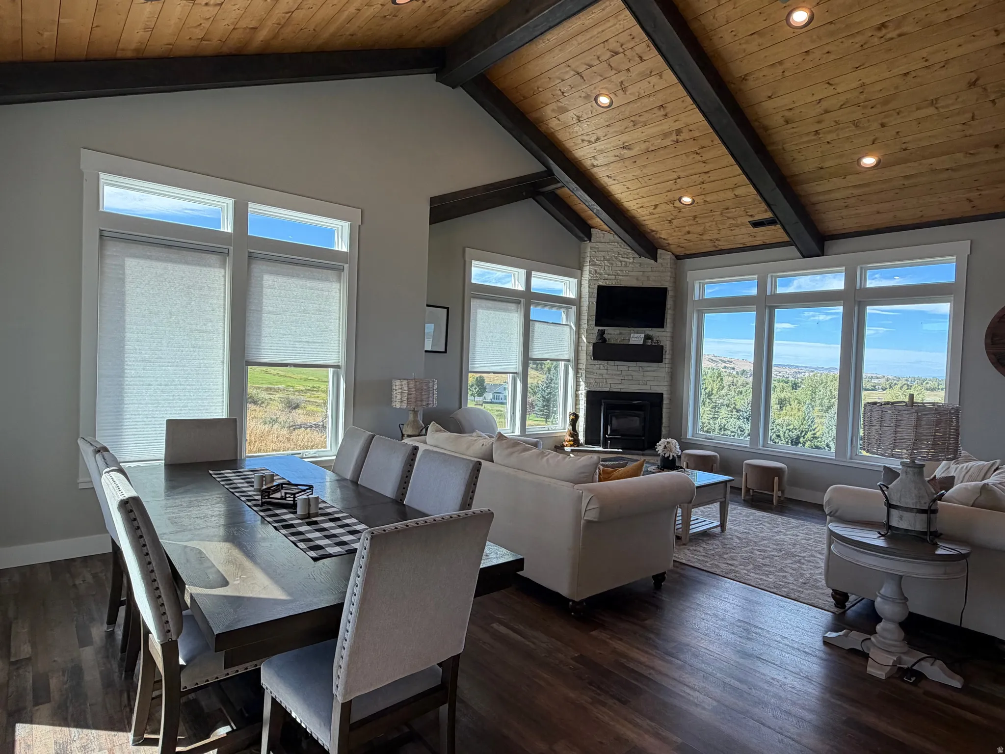Vaulted wood-paneled ceiling with exposed beams, stone-clad fireplace, large windows with roller shades, recessed lighting, and wood-finish flooring
