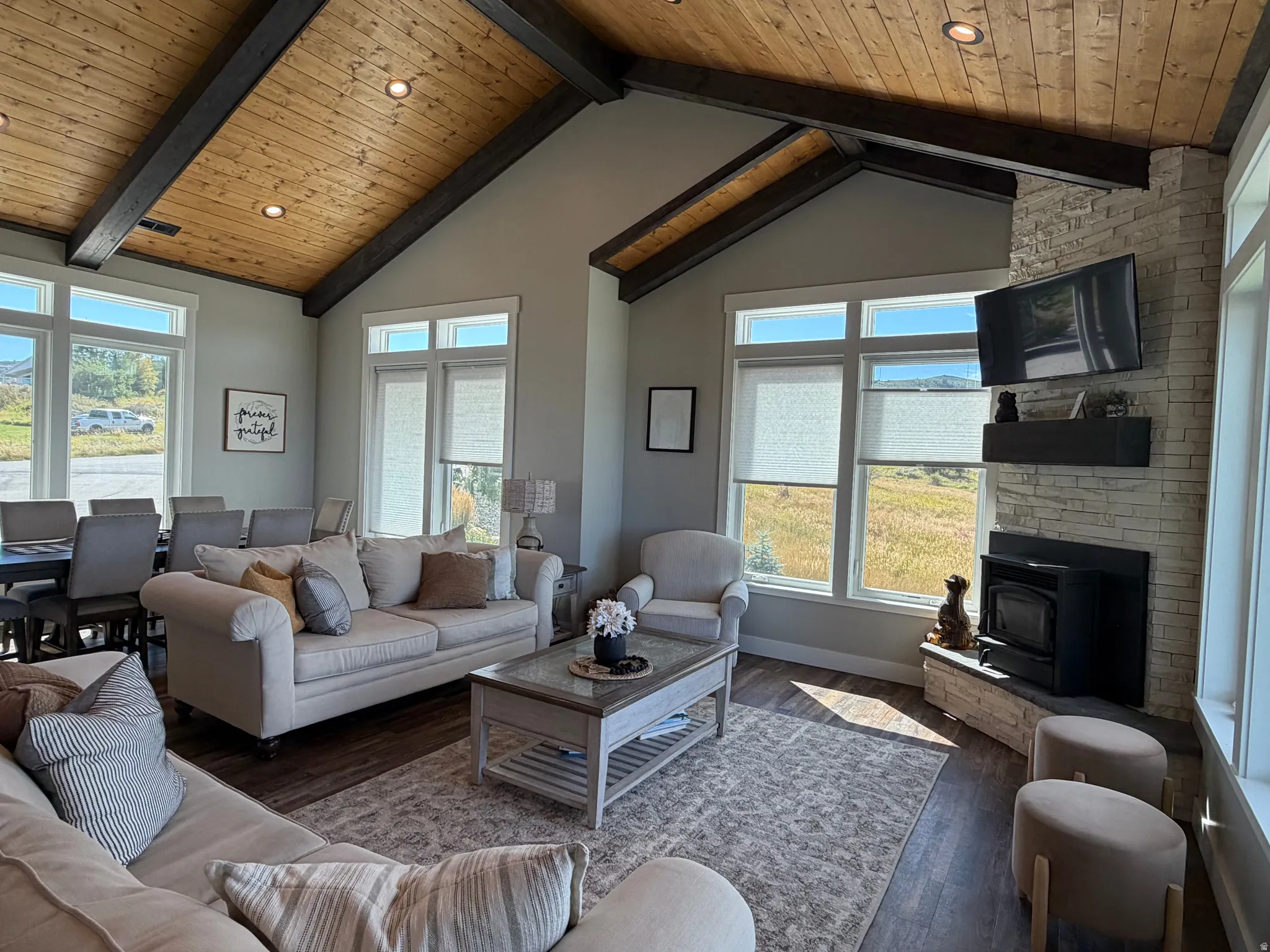 Vaulted wood-paneled ceiling with exposed beams, stone-clad fireplace with hearth, multiple large windows, and wood-finish flooring