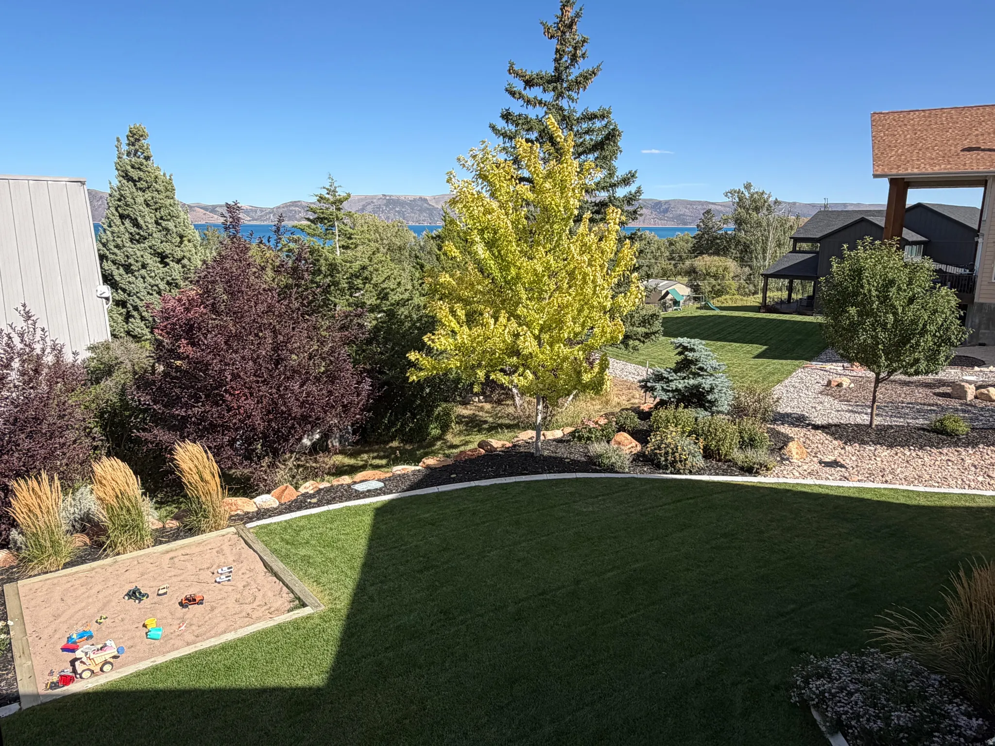 Expansive green lawn, mature trees, landscaped garden beds, sandbox, and distant body of water