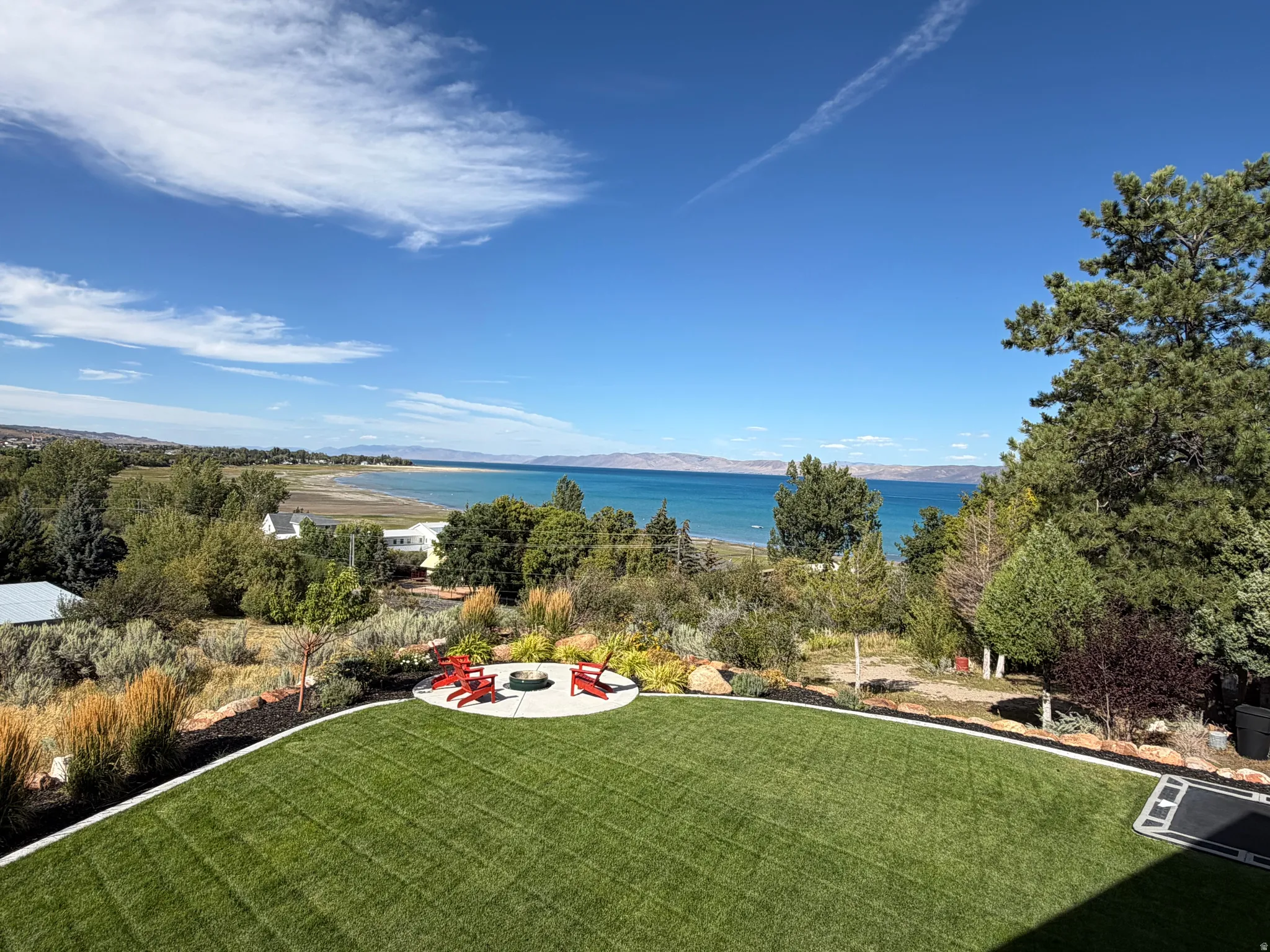 Expansive lawn with a concrete fire pit area, surrounded by mature landscaping, and featuring a distant lake and mountain backdrop