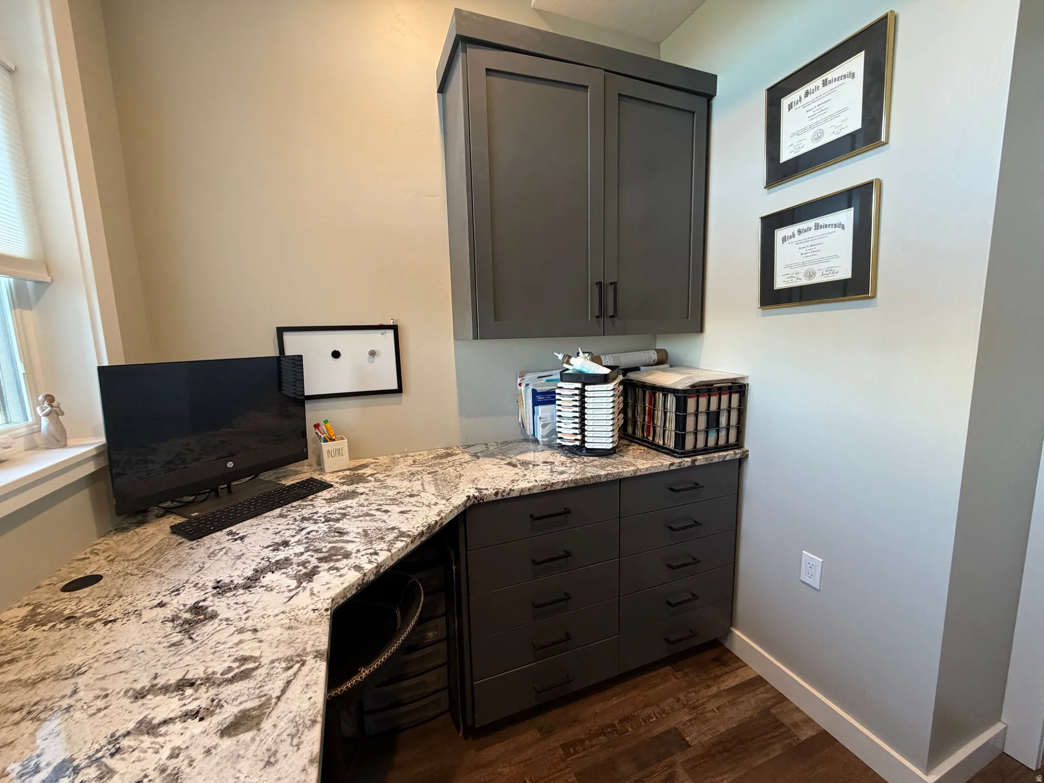 Built-in L-shaped desk with a stone countertop, featuring dark cabinetry, a built-in upper cabinet, and wood-finish flooring