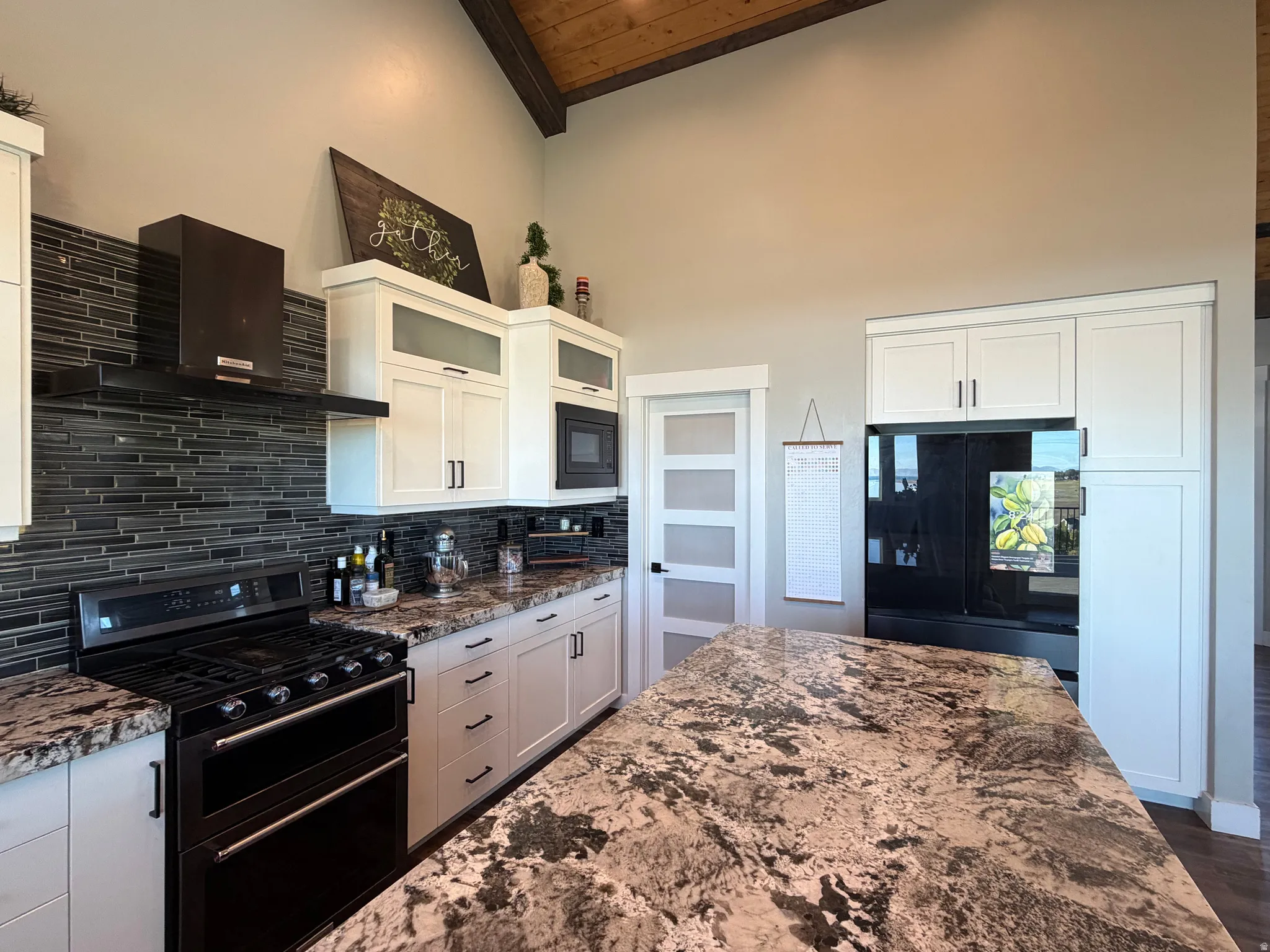 Vaulted ceiling with exposed wood beams, white shaker cabinetry with dark hardware, black tile backsplash, stainless steel range with matching hood, and a kitchen island with stone countertop
