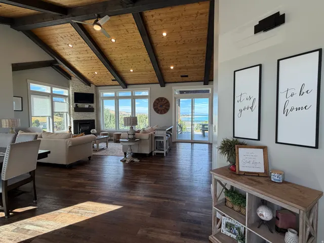 Vaulted wood-plank ceiling with exposed beams, wood-finish flooring, white brick-clad fireplace, large windows, and sliding glass doors to outdoor space