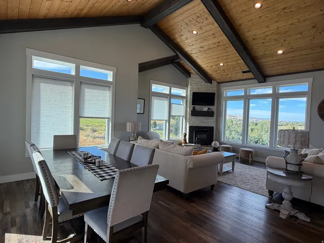 Vaulted wood-paneled ceiling with exposed beams, stone-clad fireplace, large windows with roller shades, recessed lighting, and wood-finish flooring