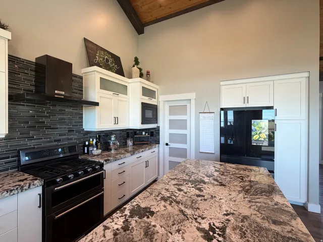 Vaulted ceiling with exposed wood beams, white shaker cabinetry with dark hardware, black tile backsplash, stainless steel range with matching hood, and a kitchen island with stone countertop