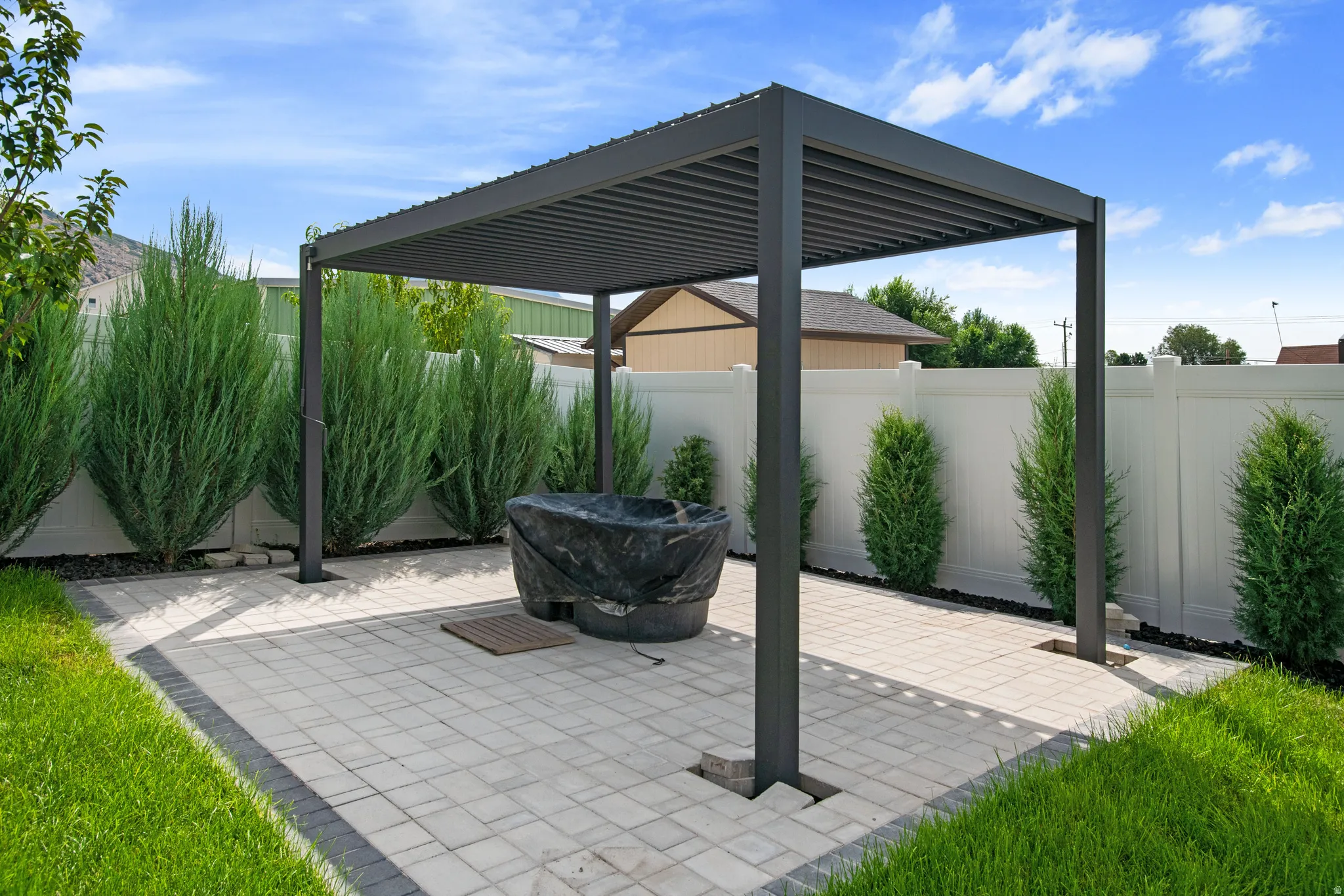 Paver patio with a dark-framed pergola, surrounded by a white privacy fence and manicured evergreen shrubs
