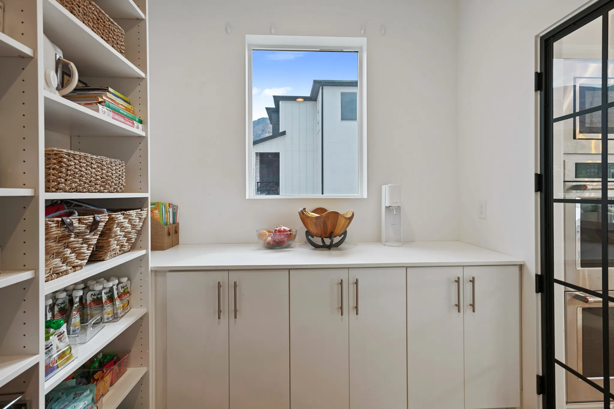 Built-in shelving, white cabinetry with silver hardware, white countertop, and a black-framed glass door