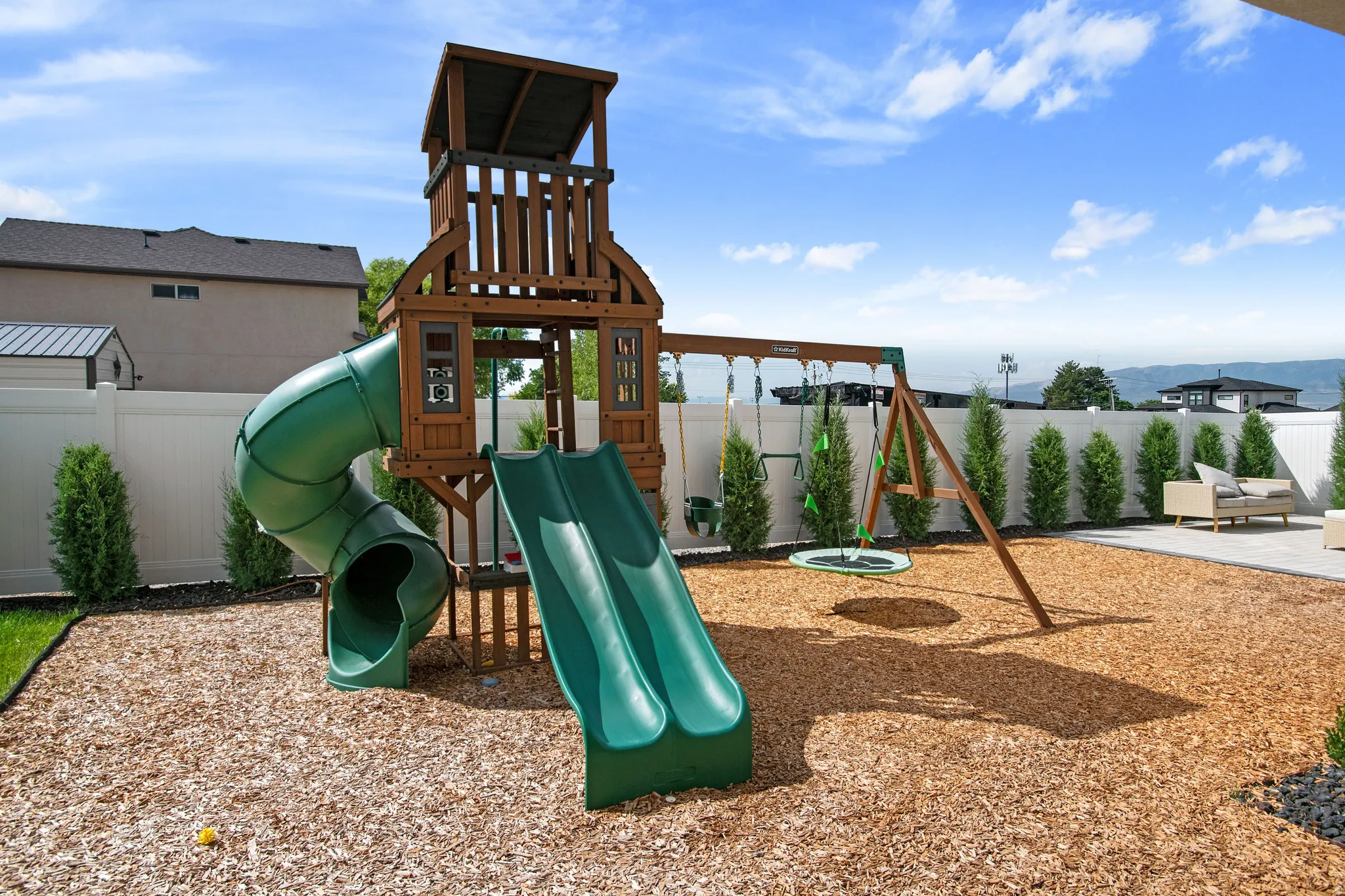 Expansive backyard with a wood chip surface, white privacy fencing, and a built-in play structure featuring slides and swings
