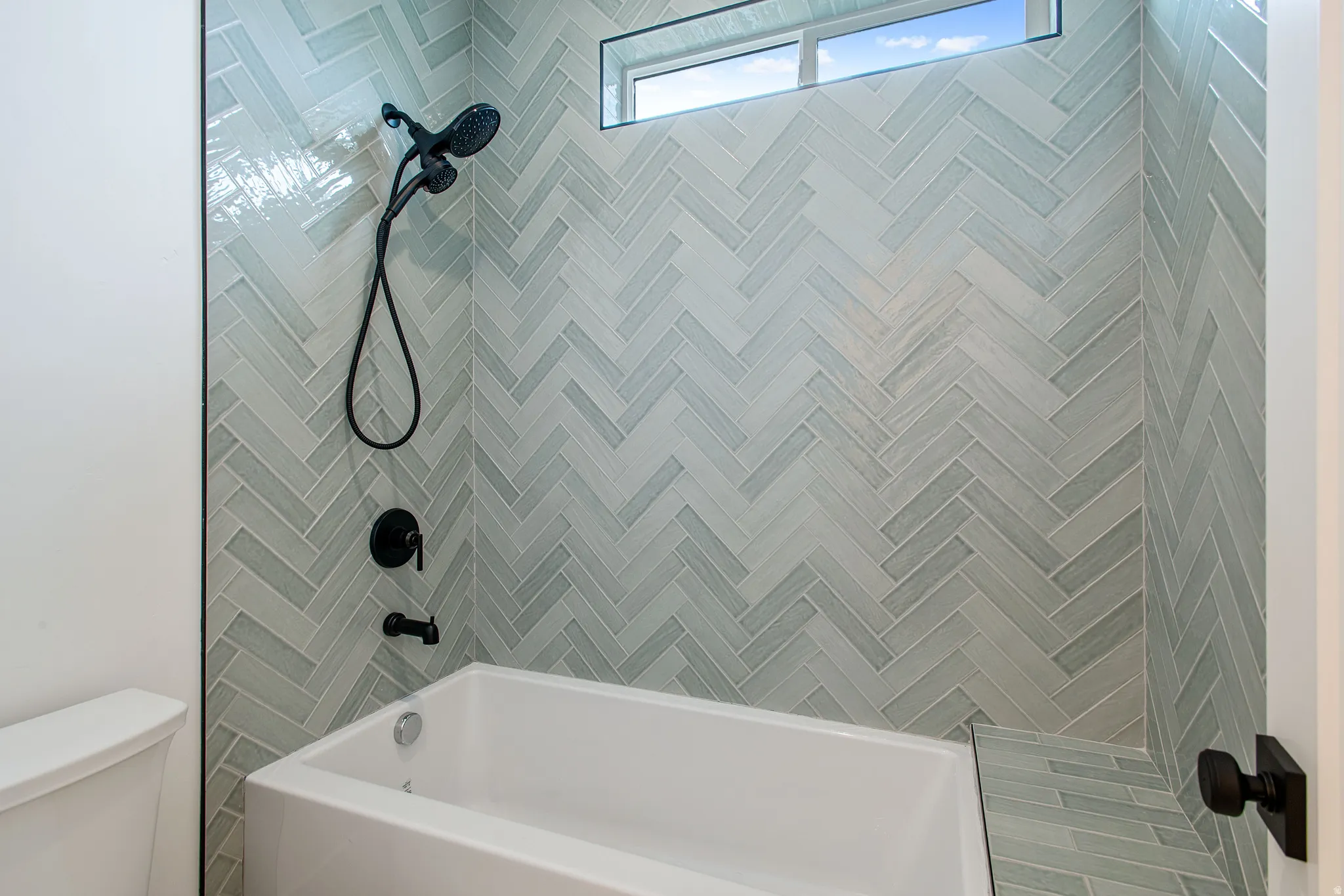 Bathtub with herringbone tile surround, matte black shower fixtures, and transom window