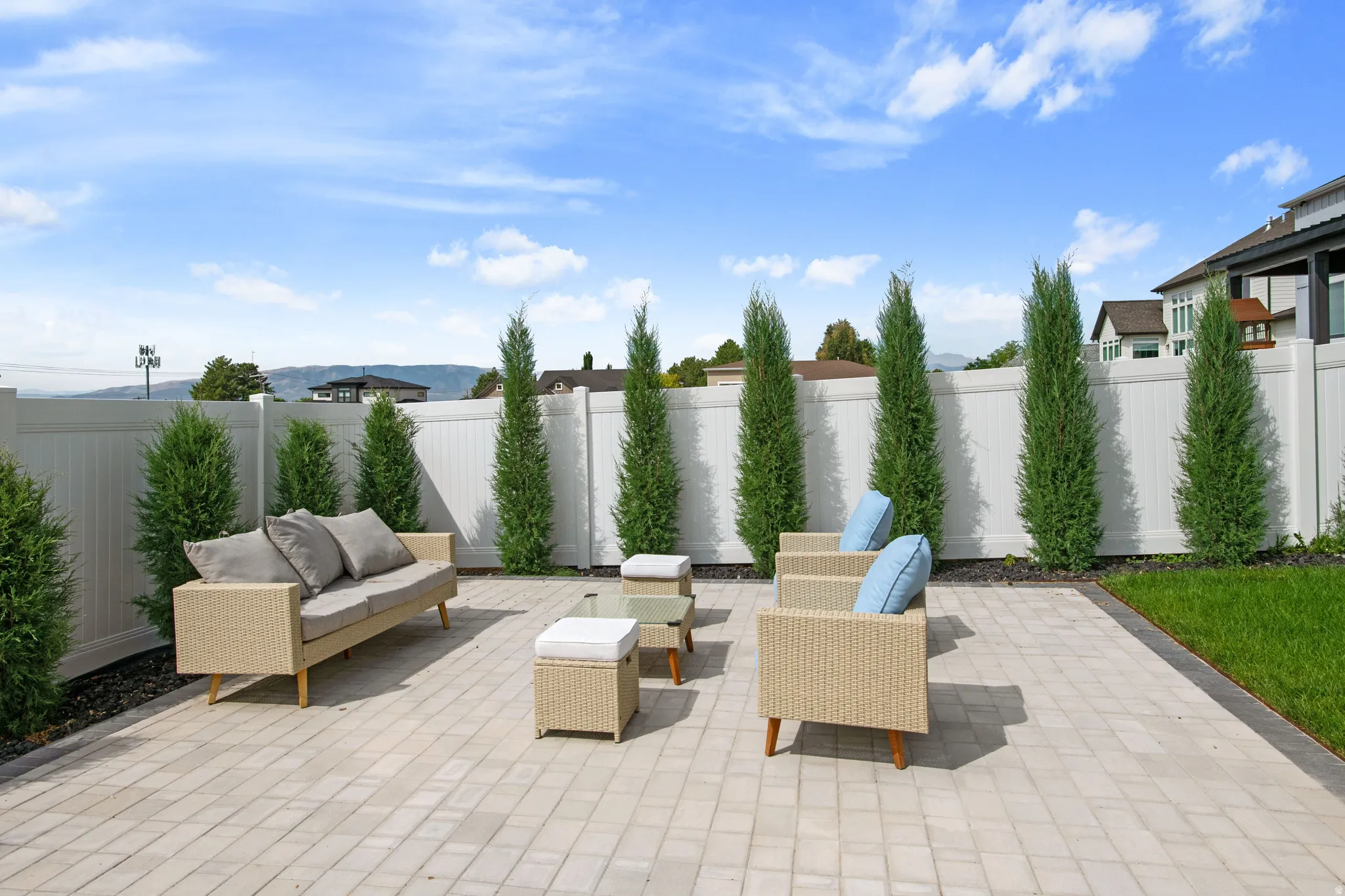 Paver patio with a white privacy fence, bordered by manicured evergreen trees and a strip of lawn