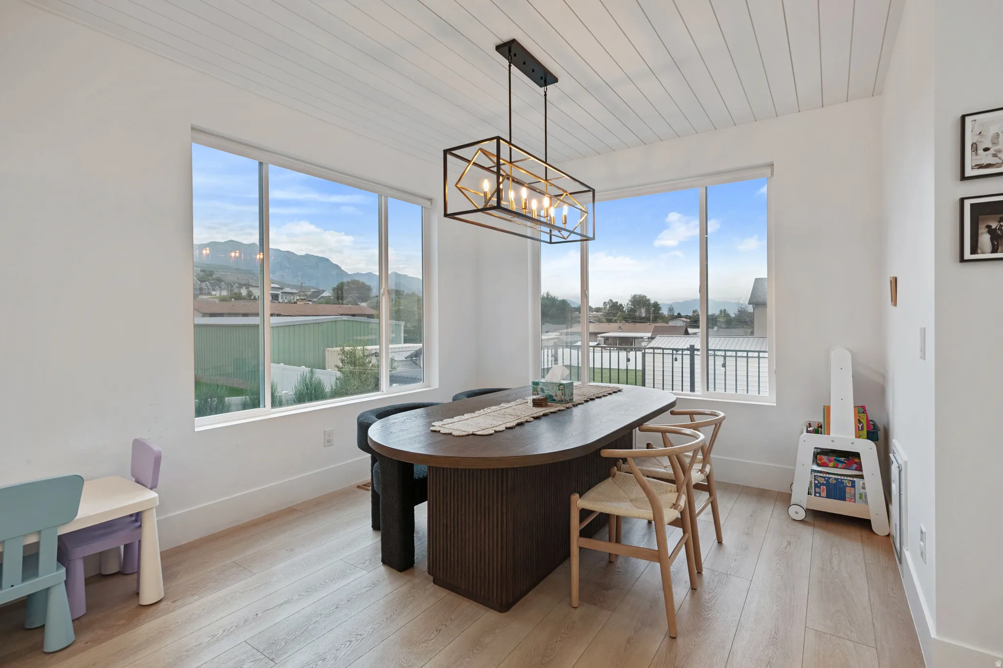 Wide plank wood-finish flooring, white shiplap ceiling, contemporary linear chandelier, expansive window openings, and white wall finishes