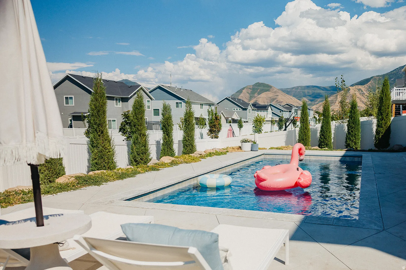 Outdoor swimming pool with concrete surround, bordered by a white fence and mature evergreen trees