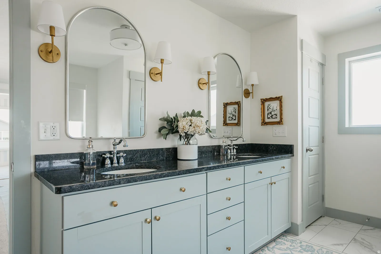 Dual arched mirrors, dual vanity with light blue cabinetry, dark stone countertops, brass wall sconces, and tiled flooring