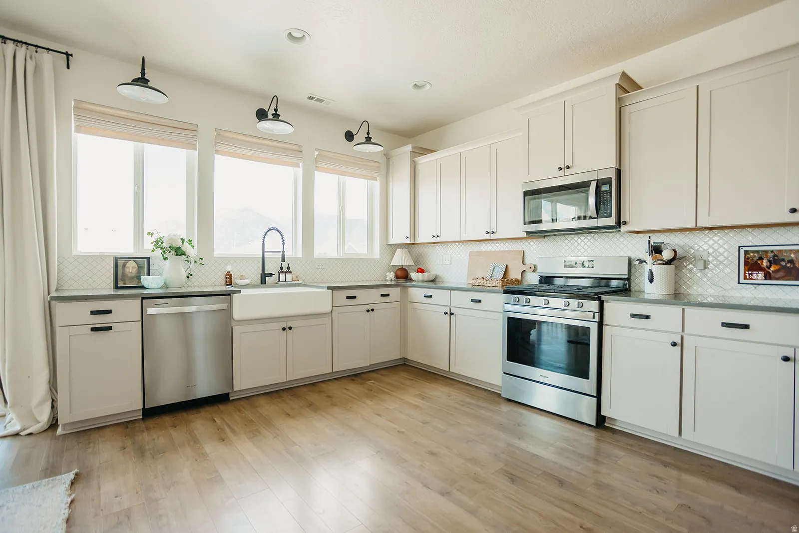 Expansive kitchen featuring wood-finish flooring, white shaker cabinetry, stainless steel appliances, a white farmhouse sink, and a white geometric tile backsplash