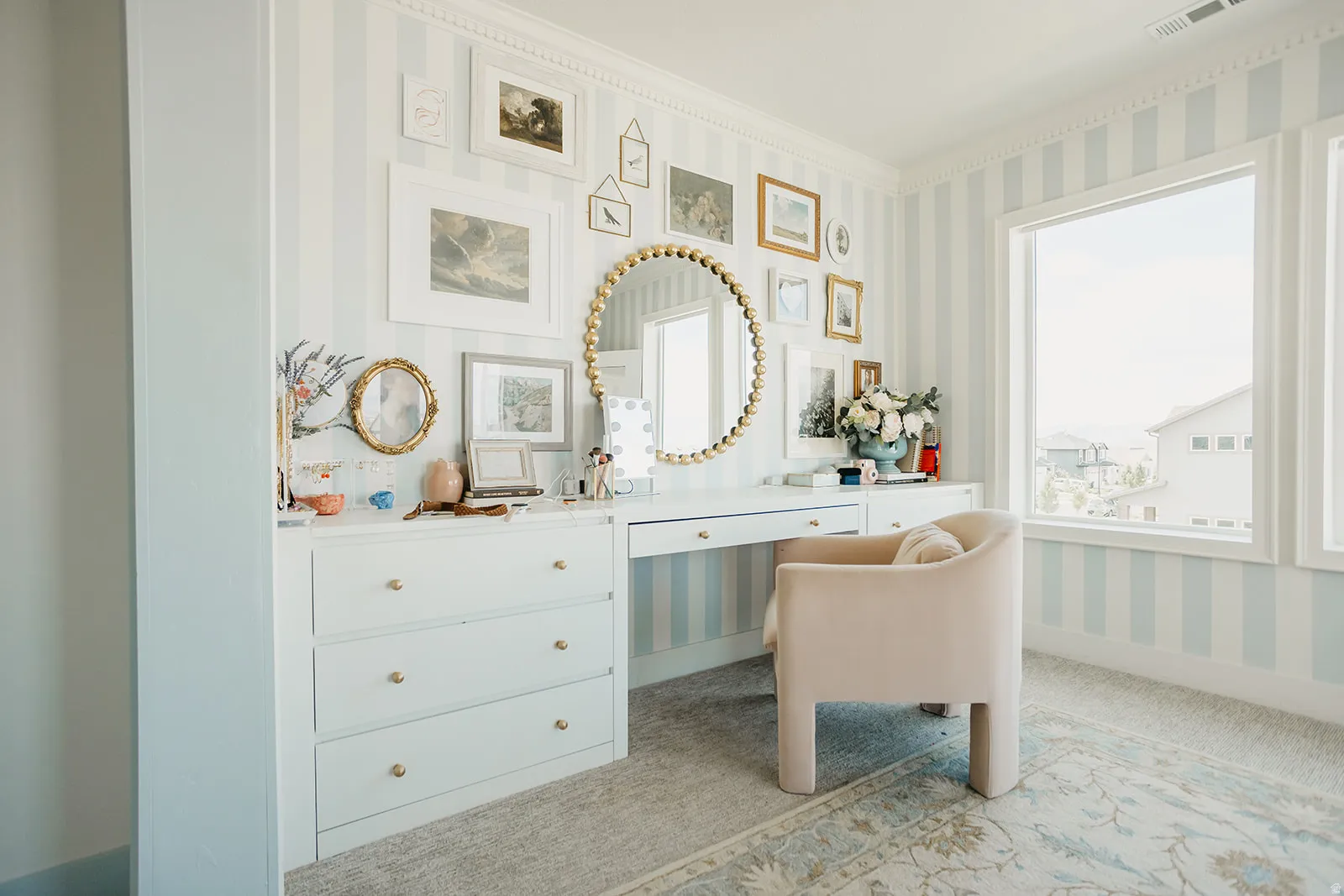 Built-in white vanity with gold hardware, broad windows, decorative wall molding, and striped wall covering