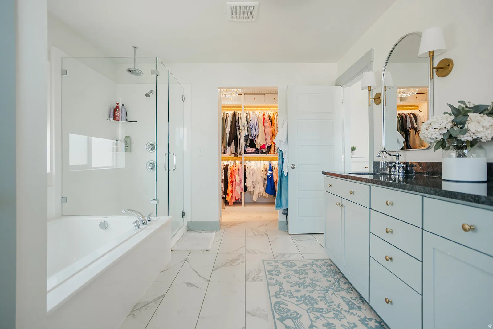 Soaking tub, glass-enclosed shower with rainhead, double vanity with integrated sink and dark countertop, large walk-in closet with built-in shelving, and large format floor tile