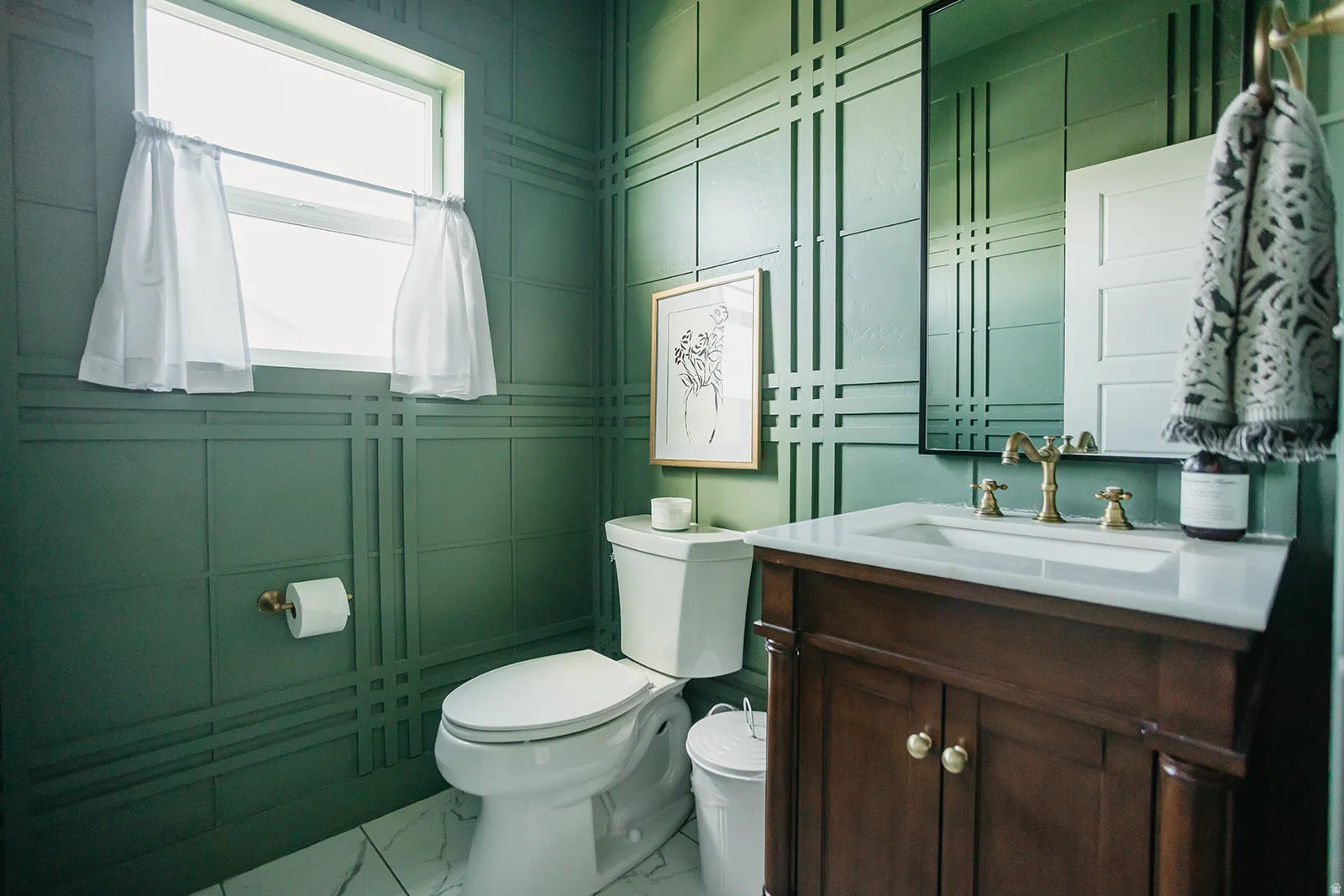Green paneled walls with a window, a toilet, a vanity with a white countertop, and a framed mirror