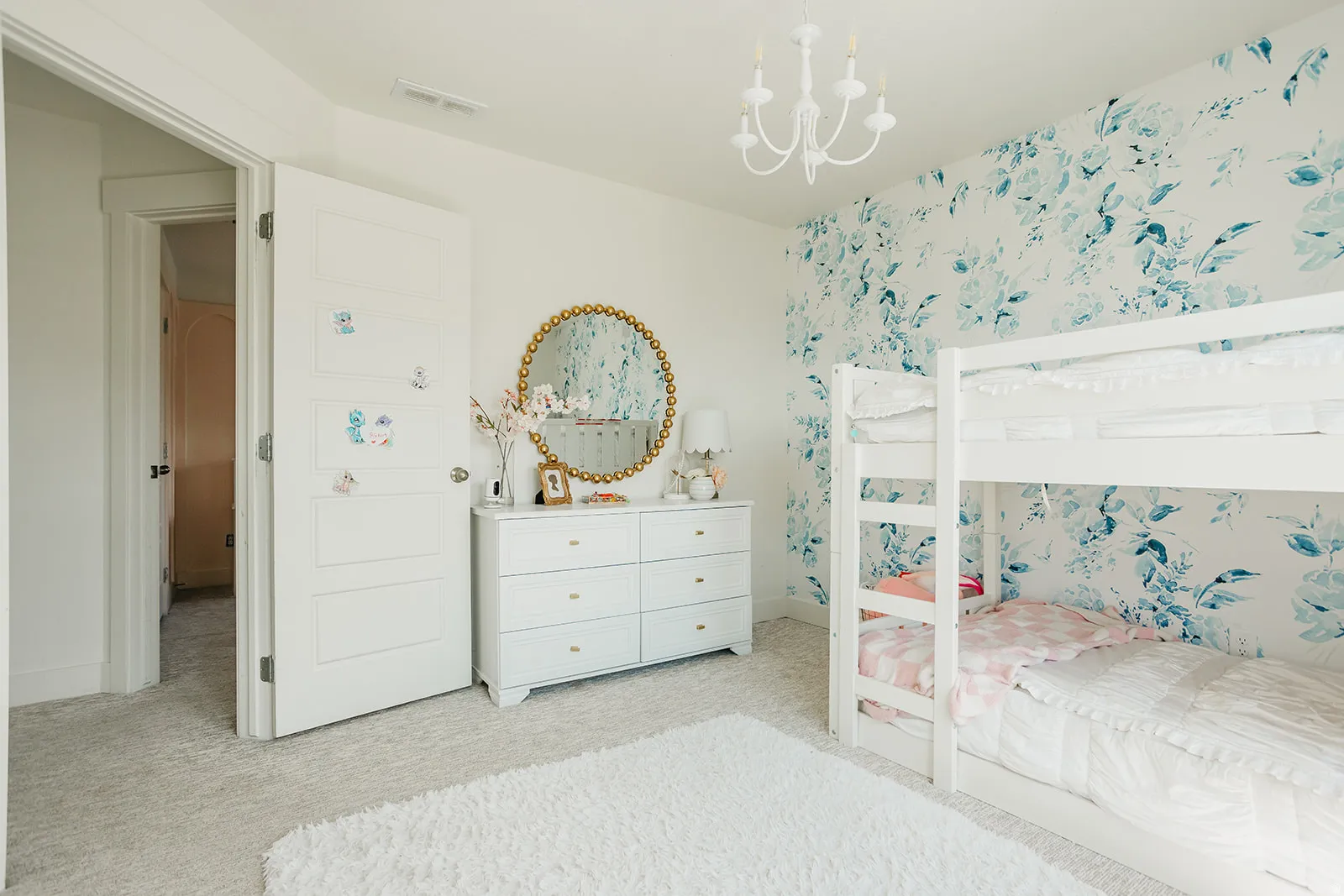 Bright, light-filled bedroom featuring white built-in bunk beds with integrated storage, white six-drawer dresser with round mirror accent, white ceiling chandelier, neutral carpeting, and decorative floral wallpaper on accent wall