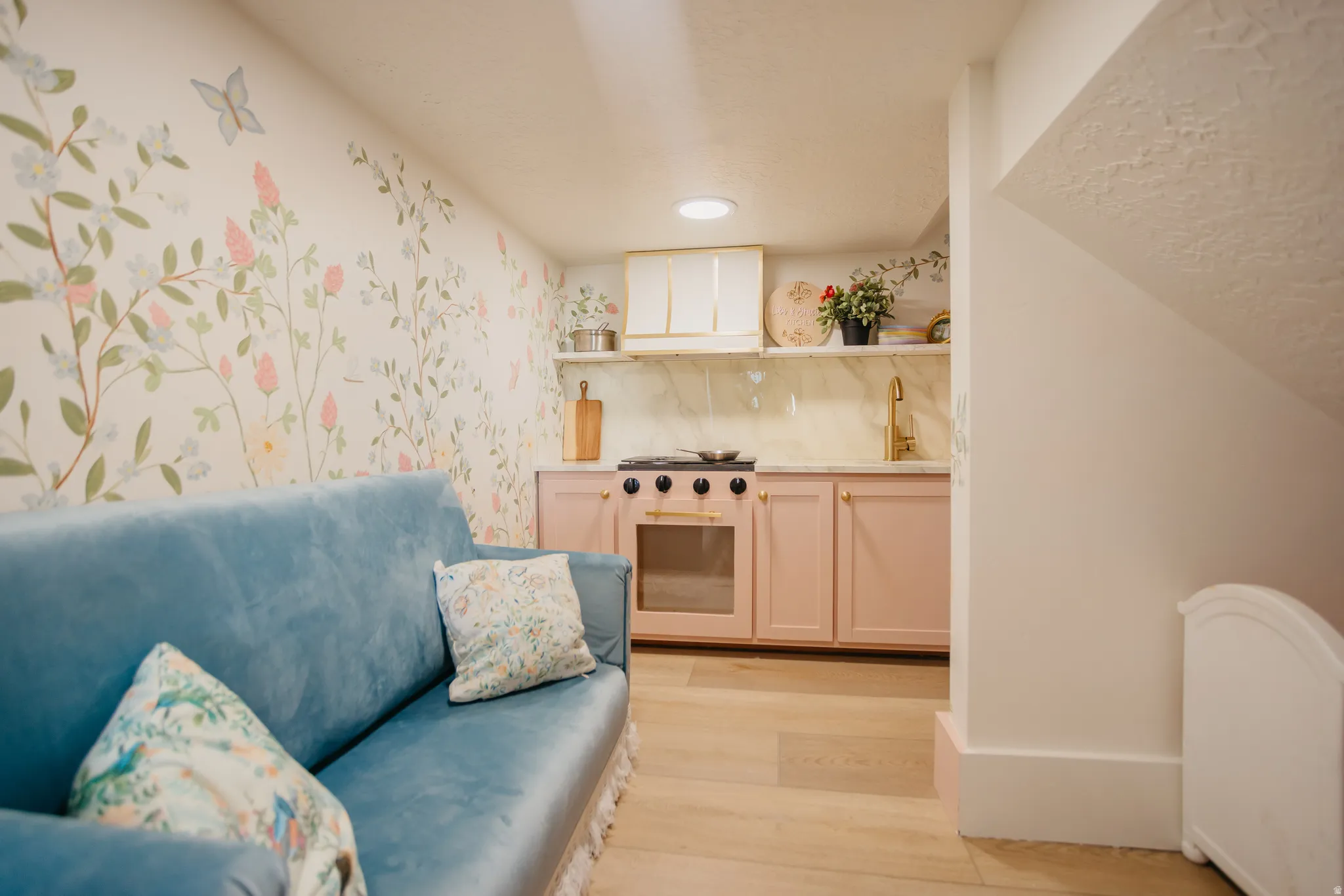 Wallpapered accent wall, wood-finish flooring, gold-tone faucet, pink cabinetry, and ceiling-mounted lighting