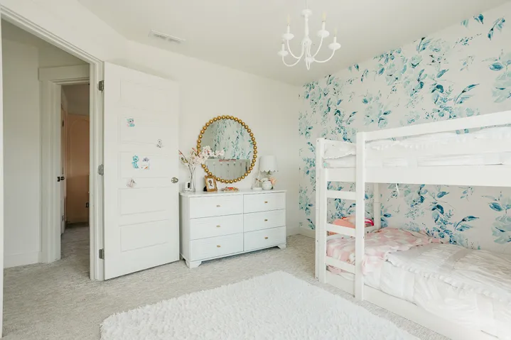 Bright, light-filled bedroom featuring white built-in bunk beds with integrated storage, white six-drawer dresser with round mirror accent, white ceiling chandelier, neutral carpeting, and decorative floral wallpaper on accent wall