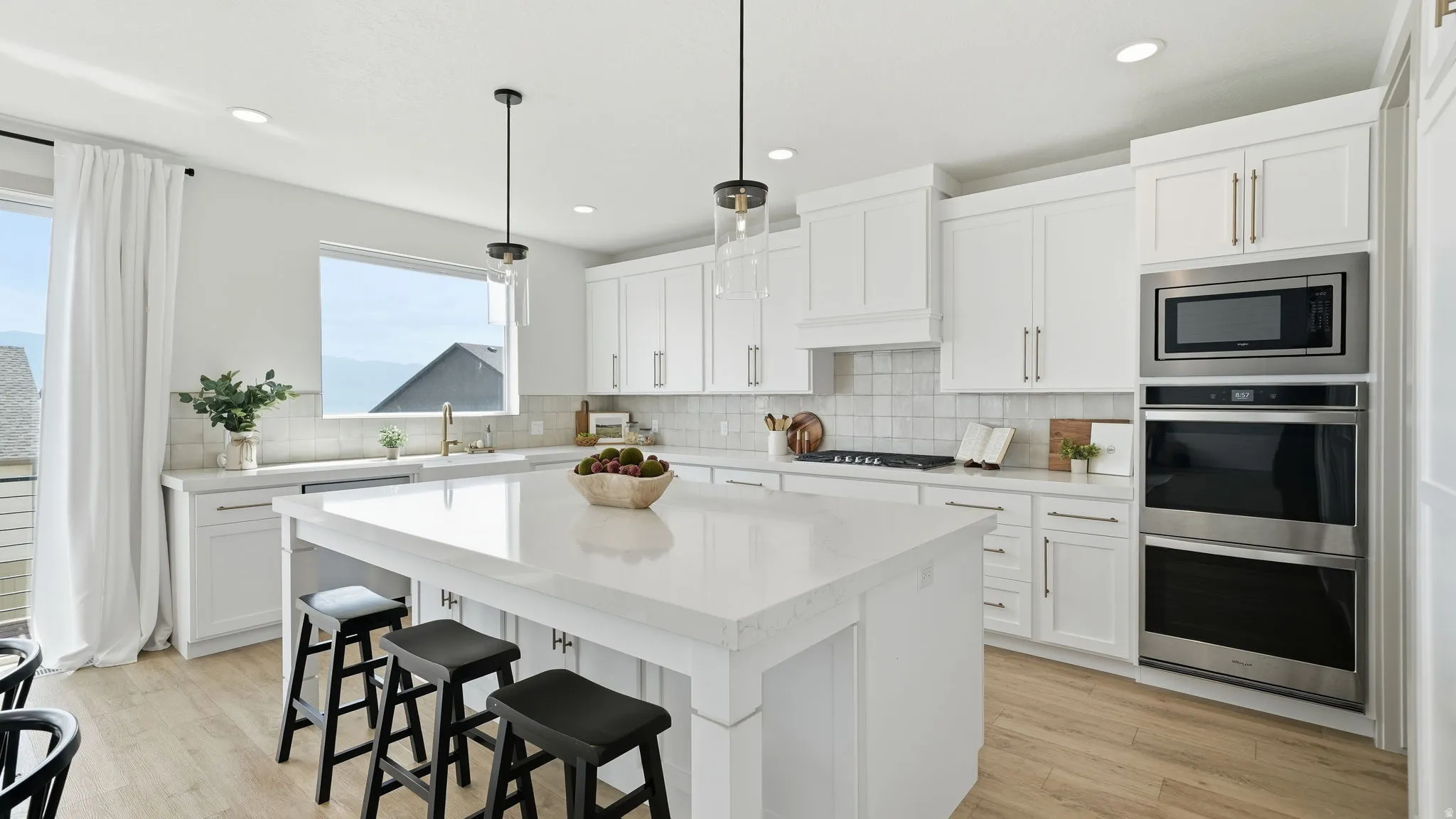 White shaker cabinetry, light-toned wood-finish flooring, recessed lighting, and stainless steel appliances including a built-in microwave and double ovens
