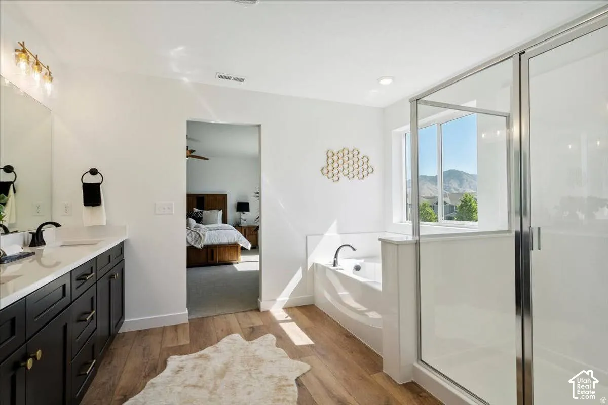 Dual vanity with dark cabinetry, wood-finish flooring, built-in bathtub, glass-enclosed shower, and large window
